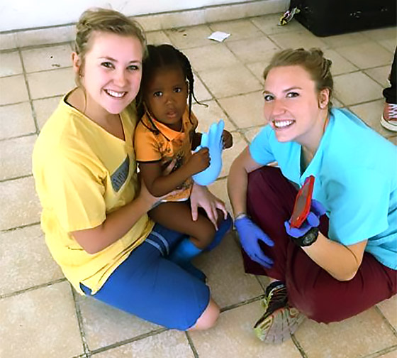 Two students pose with a child during a trip to Haiti to provide medical services. The child is holding a glove while one student holds a cell phone with a video for the child to watch.