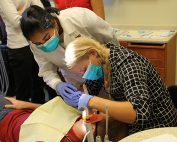 A physician assistant student gives an oral health exam to a patient as other students observe. A faculty advisor gives instructions. Both are wearing a mask and one is giving an injection.